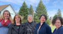 Image shows five women standing shoulder-to-shoulder with coniferous trees in the background. 