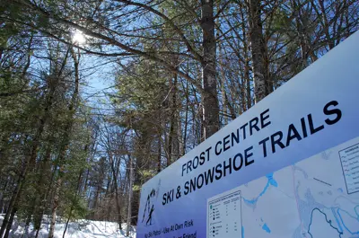 Image shows a sign for the Frost Centre Ski Trails with snow and blue sky in the background.