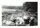 Image shows a black and white photo of a group of people eating lunch on a beach strewn with logs. 